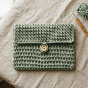 May include: A sage green rectangular crochet pouch featuring a textured stitch pattern and a single light brown wooden button closure at the center of the flap. A matching sage green yarn ball and a wooden crochet hook are positioned nearby.