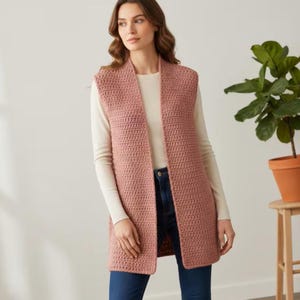 May include: A sleeveless, open-front crochet vest in a dusty rose color, worn over a long-sleeved cream-colored top and dark blue denim jeans. The vest features a consistent, textured stitch pattern and falls to the mid-thigh area.