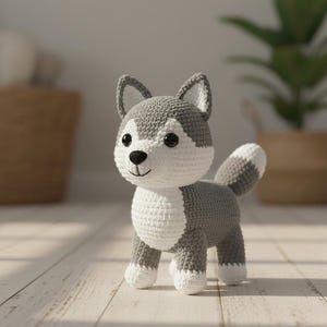 Pode incluir: Um brinquedo de cachorro husky de crochê em cinza e branco. O animal de pelúcia feito à mão tem peito, patas e rosto brancos, com detalhes cinza no corpo, orelhas e cauda. O cachorro tem olhos pretos e uma expressão sorridente.