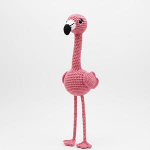May include: A handmade pink crochet flamingo with a long neck, black and white beak, and small, round wings. The flamingo has long, thin legs and small feet. The toy is made of a soft, textured yarn and stands on a white background.