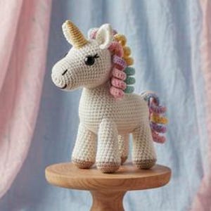 May include: A crocheted unicorn toy with a cream-colored body, a golden horn, and a multicolored mane and tail. The unicorn stands on a small wooden pedestal against a backdrop of pink and blue fabric.