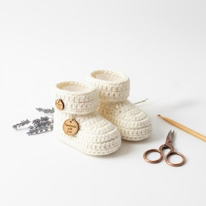 May include: A pair of cream-colored, crocheted baby booties with a wooden tag that says "Handmade with Love." The booties are next to a pair of small scissors and a wooden crochet hook. A sprig of lavender is also present.