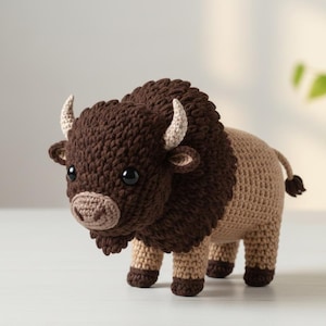 May include: A handmade crochet bison toy. The bison has a dark brown head and mane, tan body, and small cream-colored horns. The toy has black safety eyes and is standing on four legs.