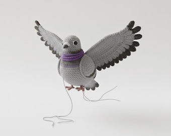 May include: A crocheted grey pigeon with outstretched wings, a purple scarf, and black wingtips. The handmade bird has a detailed texture and is suspended by thin threads, giving the impression of flight. A unique, handcrafted decorative item.