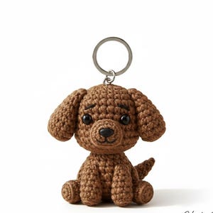 Op de afbeelding: Een bruine gehaakte honden sleutelhanger met een zilveren sleutelring. De hond heeft grote, zwarte ogen en een kleine zwarte neus. De hond zit met zijn oren naar beneden. De sleutelhanger is gemaakt van garen.