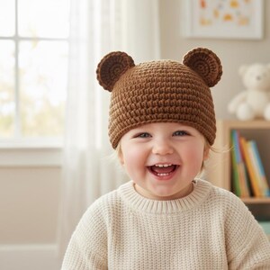 May include: A smiling child wearing a brown crochet bear hat with two round ears. The hat is made of a textured yarn. The child is wearing a cream-colored sweater.