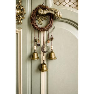 May include: A decorative door hanging with a woven wreath and three brass bells. It features small glass bottles, wooden beads, and a crescent moon charm. The door is painted a muted green. The door handle is ornate.