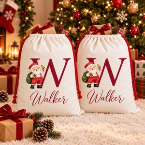 Pode incluir: Dois sacos de presente de tecido off-white com cordão, apresentando uma letra W grande em vermelho escuro, um gráfico do Papai Noel segurando um sino e um saco verde, e o nome Walker impresso em letra cursiva vermelha. Cada saco tem cordões vermelhos.