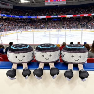 Puede incluir: Tres juguetes de peluche con forma de disco de hockey con caras sonrientes y piernas y patines adjuntos. Los discos son grises con detalles en negro y turquesa. Cada disco tiene las letras "OK". Los juguetes se muestran frente a una pista de hockey.