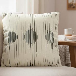 Op de afbeelding: Vierkant decoratief kussen met een crèmekleurige achtergrond en een geometrisch patroon. Het patroon bevat verticale lijnen en drie ruitvormige ontwerpen in grijstinten en zwart. Het kussen ligt op een lichtgekleurde bank, ongeveer 40 cm x 40 cm.
