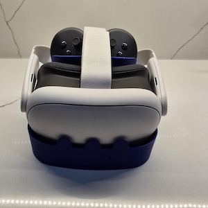 May include: A white and blue VR headset with two black controllers. The headset has a white strap and a blue base. The controllers are black with gray buttons. The headset is designed for virtual reality gaming and entertainment.