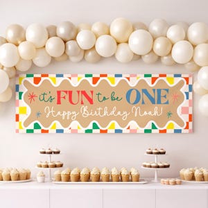 May include: A birthday banner with the text "it's FUN to be ONE Happy Birthday Noah!" is displayed above a table with cupcakes. The banner has a brown background with a colourful border and text in red, blue, and green.