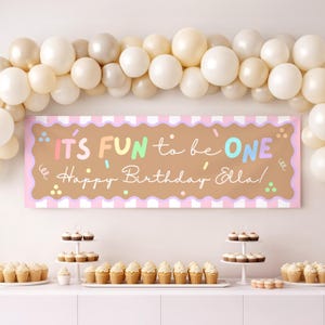 Könnte beinhalten: Ein Geburtstagsbanner mit dem Text "IT'S FUN to be ONE Happy Birthday Ella!" in Pastellfarben, mit einem rosa-weiß gestreiften Rand. Das Banner ist mit bunten Konfetti verziert und von einem Ballonbogen und Cupcakes umgeben.