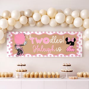 以下が含まれることがあります： 「Oh Twodles! Haleigh is 2」のピンクと白の文字が入った誕生日バナー。ミニーマウスのグラフィックが特徴です。バナーは茶色の背景とピンクのハートの装飾が施されています。風船のアーチとカップケーキも表示されています。