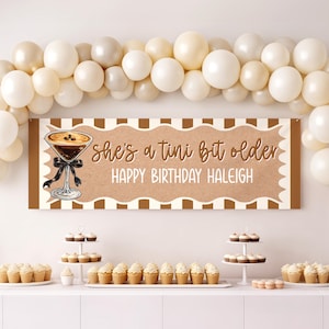May include: A rectangular birthday banner with a brown and white scalloped border, featuring a martini glass illustration with a black bow and the text: she's a tini bit older HAPPY BIRTHDAY HALEIGH. The banner is displayed under white balloons.