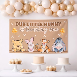 Puede incluir: Una pancarta marrón con el texto "OUR LITTLE HUNNY is turning one" e ilustraciones de Winnie the Pooh, Piglet, Tigger y Eeyore. La pancarta está decorada con panales y puntos. Globos en blanco, beige y plata están arriba.