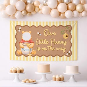 May include: A rectangular banner with a light yellow striped background and a brown scalloped center featuring a cartoon bear holding a honey pot, bees, a beehive, and the text Our Little Hunny is on the way, topped with a balloon arch.