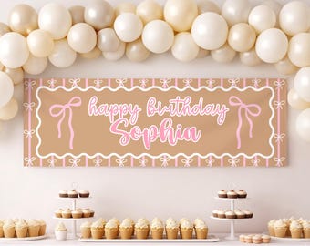 May include: A rectangular tan birthday banner featuring a white scalloped border, pink vertical stripes, and pink bow illustrations. The text reads "happy birthday Sophia" in white script. The banner is displayed beneath a large arch of cream balloons.
