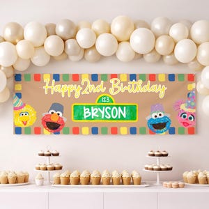 May include: A birthday banner with the text "Happy 2nd Birthday" and the name "Bryson". The banner features cartoon characters and colorful squares. White balloons are arranged above the banner, and cupcakes are displayed on a table.