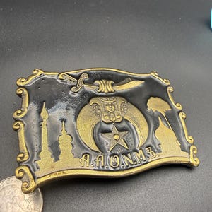 May include: Rectangular metal belt buckle with a black background and gold-colored accents. The buckle features a sword, a star, and the letters "A.A.O.N.M.S." along with other symbols. The edges have a decorative border.