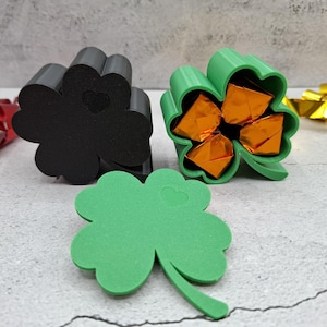 May include: Three-leaf clover-shaped containers in black and green, with a green clover cutout. The green container holds wrapped orange sweets. A heart is cut out of the clovers. Red and gold ribbon are in the background.