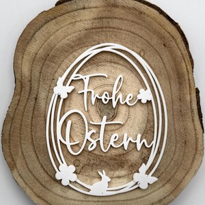 Puede incluir: Decoración de Pascua de madera con las palabras "Frohe Ostern" en escritura blanca, rodeada por un marco ovalado blanco adornado con motivos florales y de conejos. La rodaja de madera es de color marrón claro natural.