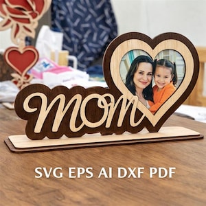 May include: A wooden photo frame with the word "Mom" in a cursive font. The frame has a heart-shaped cutout holding a photo of a woman and a child. The base is rectangular, and the frame is made of light and dark wood.