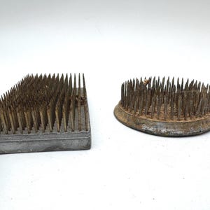 May include: A rectangular metal flower frog with a grid of upright spikes and a circular metal flower frog with a dense arrangement of vertical spikes. Both items feature a weathered, aged finish with visible oxidation on the metal surfaces.