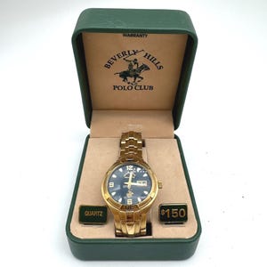 May include: A gold-tone analog wristwatch with a dark face, housed in a green presentation box. The interior lid features the Beverly Hills Polo Club logo and text. Two small rectangular plaques labeled Quartz and 150 are placed beside the watch.