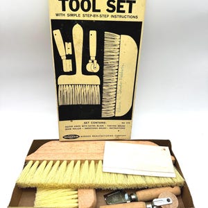 Könnte beinhalten: Ein Tapezierwerkzeug-Set der Warner Manufacturing Company in einem gelben Karton. Das Set enthält eine hölzerne Tapezierbürste mit hellgelben Borsten, eine Nahtrolle, ein Cuttermesser und einen Kleisterpinsel, alle mit Holzgriffen.