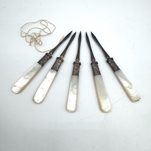 May include: Five pointed metal picks with iridescent white mother-of-pearl handles and ornate silver-toned metal ferrules. The picks are arranged in a fan shape, and a thin, light-colored string is coiled near the top of the leftmost pick.