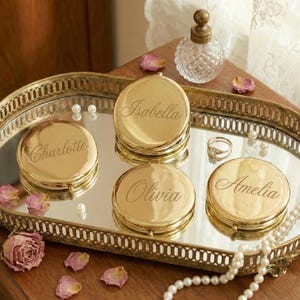 Op de afbeelding: Een goudkleurige vanity tray met een spiegeloppervlak bevat vier gepersonaliseerde compacts, elk gegraveerd met een andere naam: "Charlotte", "Isabella", "Olivia" en "Amelia". Een kleine parfumfles en een ring liggen ook op de tray, samen met parels en gedroogde rozenblaadjes.