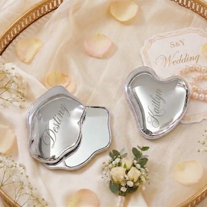 May include: Two silver compact mirrors, one petal-shaped and one heart-shaped, are displayed on a gold tray with a cream-colored cloth. The petal-shaped mirror has the name "Destiny" engraved, and the heart-shaped mirror has the name "Kaitlyn".