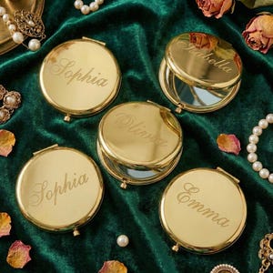 Peut inclure: Miroirs compacts dor&eacute;s avec les noms grav&eacute;s Sophia, Olivia, Emma et Isabella. Les compacts ronds s'ouvrent pour r&eacute;v&eacute;ler un miroir. Le fond est un tissu de velours vert fonc&eacute; avec des perles et des fleurs s&eacute;ch&eacute;es.