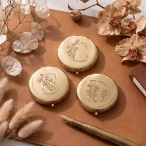 Op de afbeelding: Drie ronde, goudkleurige metalen compactspiegels, elk voorzien van een gegraveerde letter C, S of D, versierd met een delicaat bloemmotief in lijntekening. De spiegels liggen op een beige ondergrond naast een gouden vulpen en gedroogde bloemen.