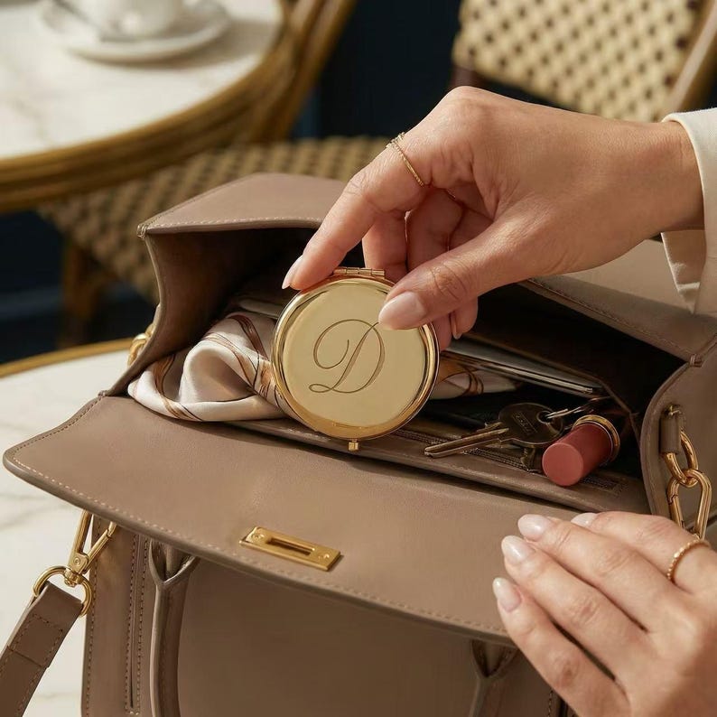 May include: A gold compact mirror with the letter "D" engraved on the front is held open, revealing a reflection. The mirror is inside a taupe handbag, along with keys, a lipstick, and a patterned scarf. The handbag has a gold clasp and chain.