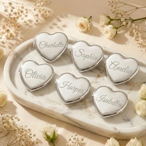 May include: Six heart-shaped, silver compact mirrors with engraved names: Charlotte, Olivia, Sophia, Harper, Amelia, and Isabella. The mirrors are arranged on a white marble tray, surrounded by white flowers.