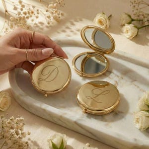 May include: Gold-colored compact mirrors with engraved initials. One mirror is open, revealing a reflective surface. The other two mirrors are closed, each displaying a different initial. The mirrors are on a marble tray, surrounded by white roses and delicate flowers.