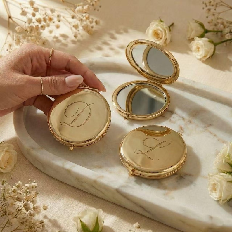 May include: Gold-colored compact mirrors with engraved initials. One mirror is open, revealing a reflective surface. The other two mirrors are closed, each displaying a different initial. The mirrors are on a marble tray, surrounded by white roses and delicate flowers.