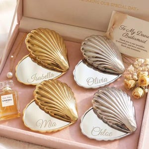 May include: A gift box with four seashell-shaped compact mirrors, two gold and two silver, each engraved with a name. A perfume bottle and a necklace with a pearl pendant are also in the box. A card reads "To My Dearest Bridesmaid."