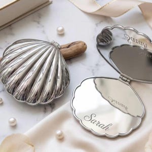 Op de afbeelding: Een zilverkleurige, schelpvormige compacte spiegel met een metallic afwerking. Eén spiegel is gesloten en toont een geribbelde buitenkant, terwijl de andere open is en een reflecterend oppervlak onthult met de naam Sarah in een cursief lettertype.