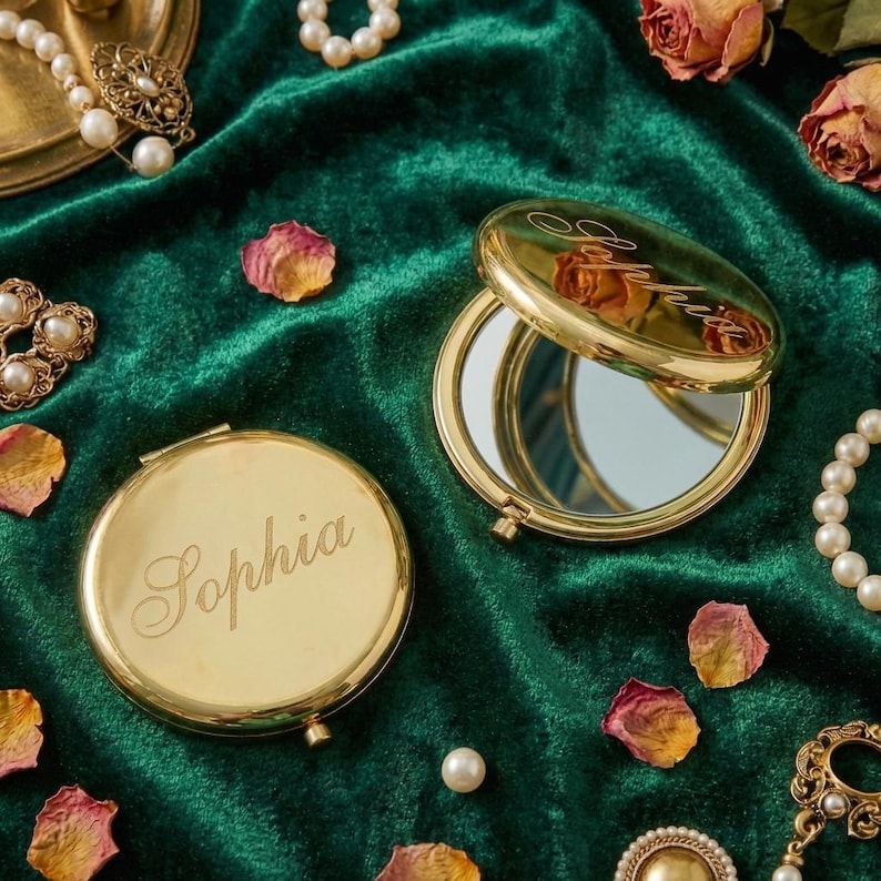 Peut inclure: Miroirs compacts dor&eacute;s avec le nom "Sophia" grav&eacute;. L'un est ouvert, r&eacute;v&eacute;lant une surface r&eacute;fl&eacute;chissante. Ils reposent sur un tissu de velours vert fonc&eacute;, entour&eacute;s de perles et de p&eacute;tales de roses s&eacute;ch&eacute;es.