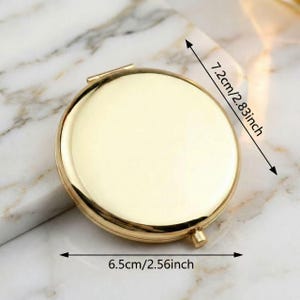 May include: A gold-colored, round compact mirror. The mirror is 2.56 inches in diameter and 2.83 inches in height. The compact is open, revealing the mirror surface. The background is a marble-like pattern.