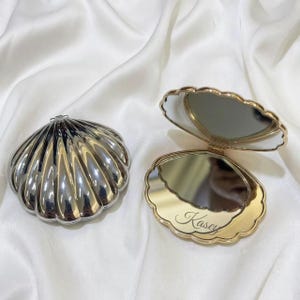 Peut inclure: Deux miroirs de poche en forme de coquillage avec une finition métallique. Un miroir est fermé, montrant un extérieur argenté avec des stries. L'autre est ouvert, révélant une surface réfléchissante et le nom Kasey gravé en cursive.