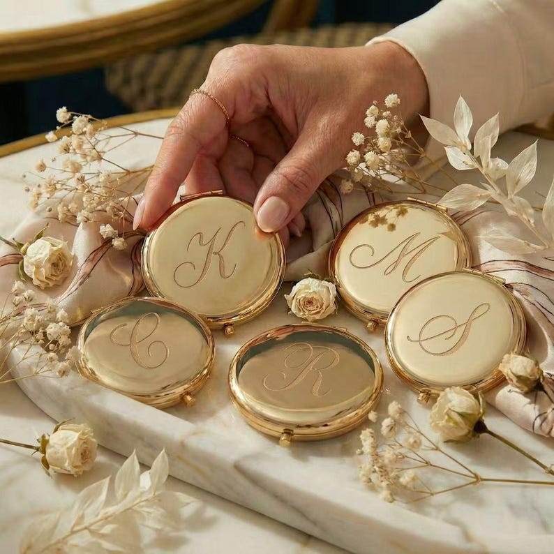 May include: Gold compact mirrors with engraved initials, including "K", "M", "S", "C", and "R". The compacts are round and sit on a marble surface, surrounded by dried flowers and a hand.