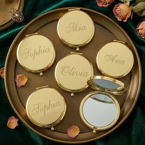 May include: Gold-colored compact mirrors with personalized names engraved on the lids. One mirror is open, revealing a reflective surface. The names Mia, Sophia, Olivia, and Ava are visible. The mirrors are arranged on a decorative tray.