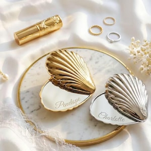Op de afbeelding: Twee schelpvormige compacte spiegels, één goudkleurig en één zilverkleurig, liggen op een marmeren dienblad. De gouden compact is gegraveerd met "Penelope", en de zilveren met "Charlotte". Een gouden lipstick, ringen en een parelketting zijn ook aanwezig.