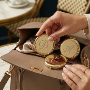 May include: Gold compact mirrors with engraved initials D, A, and E are displayed inside a taupe handbag. The mirrors are round and have a shiny finish. A silk scarf is visible inside the bag.