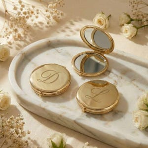 May include: Three gold-colored compact mirrors, two closed and one open, with a marble tray and white roses. The closed compacts have the letters "D" and "L" engraved on them. The open compact mirror reflects the surrounding objects.