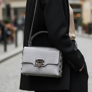 Puede incluir: Un bolso gris claro con un estampado de cocodrilo y herrajes plateados. El bolso tiene un asa superior y una correa de cadena. La mujer lleva una blazer y un pantalón negros.